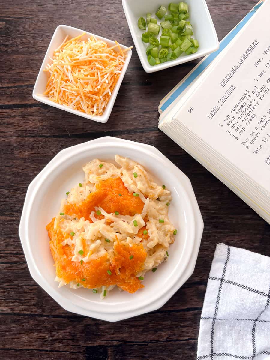 white plate with cheesy potatoes with a small dish of shredded cheese and a green onions alongside a cookbook.
