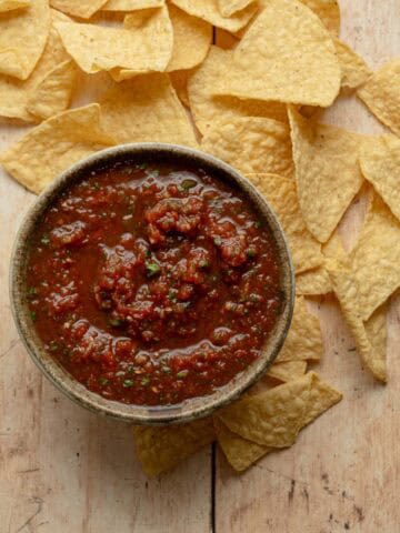 bowl of salsa surrounded by tortilla chips on a wooden board.