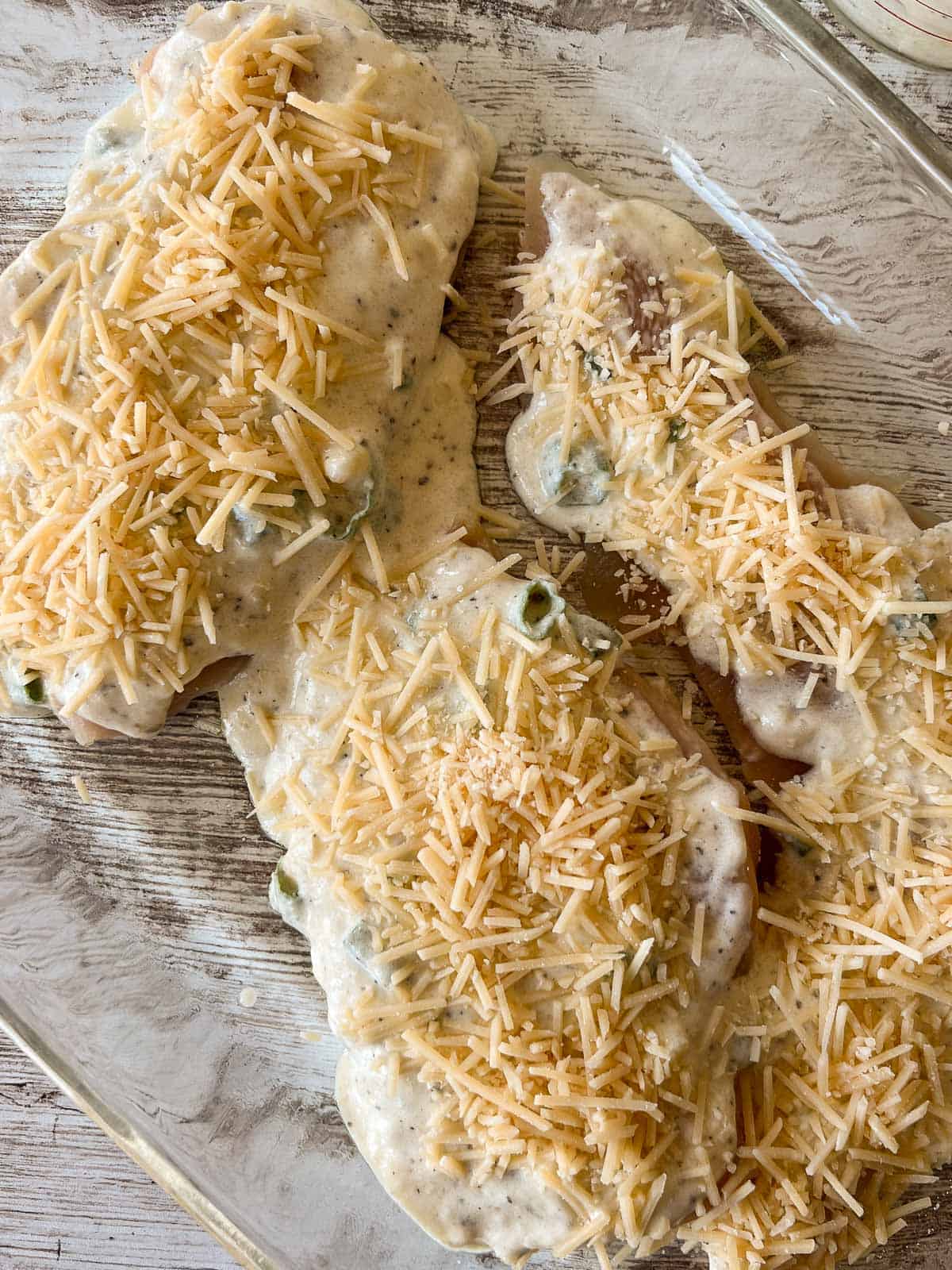 coated piece of chicken in a glass baking pan, topped with shredded cheese.