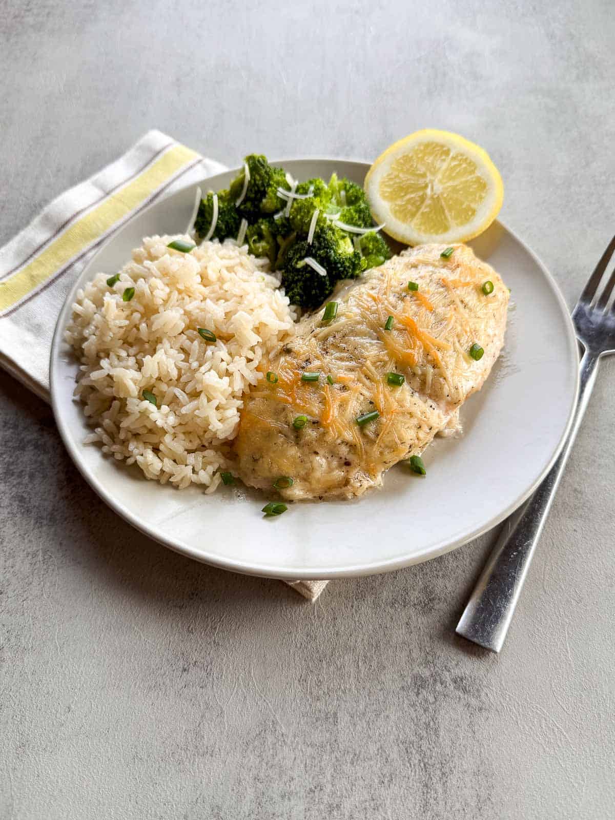 piece of baked chicken, a scoop of rice and broccoli with a lemon slice on a plate.