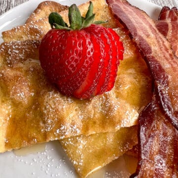 pancakes served on a white plate with a side of bacon and sliced strawberry.