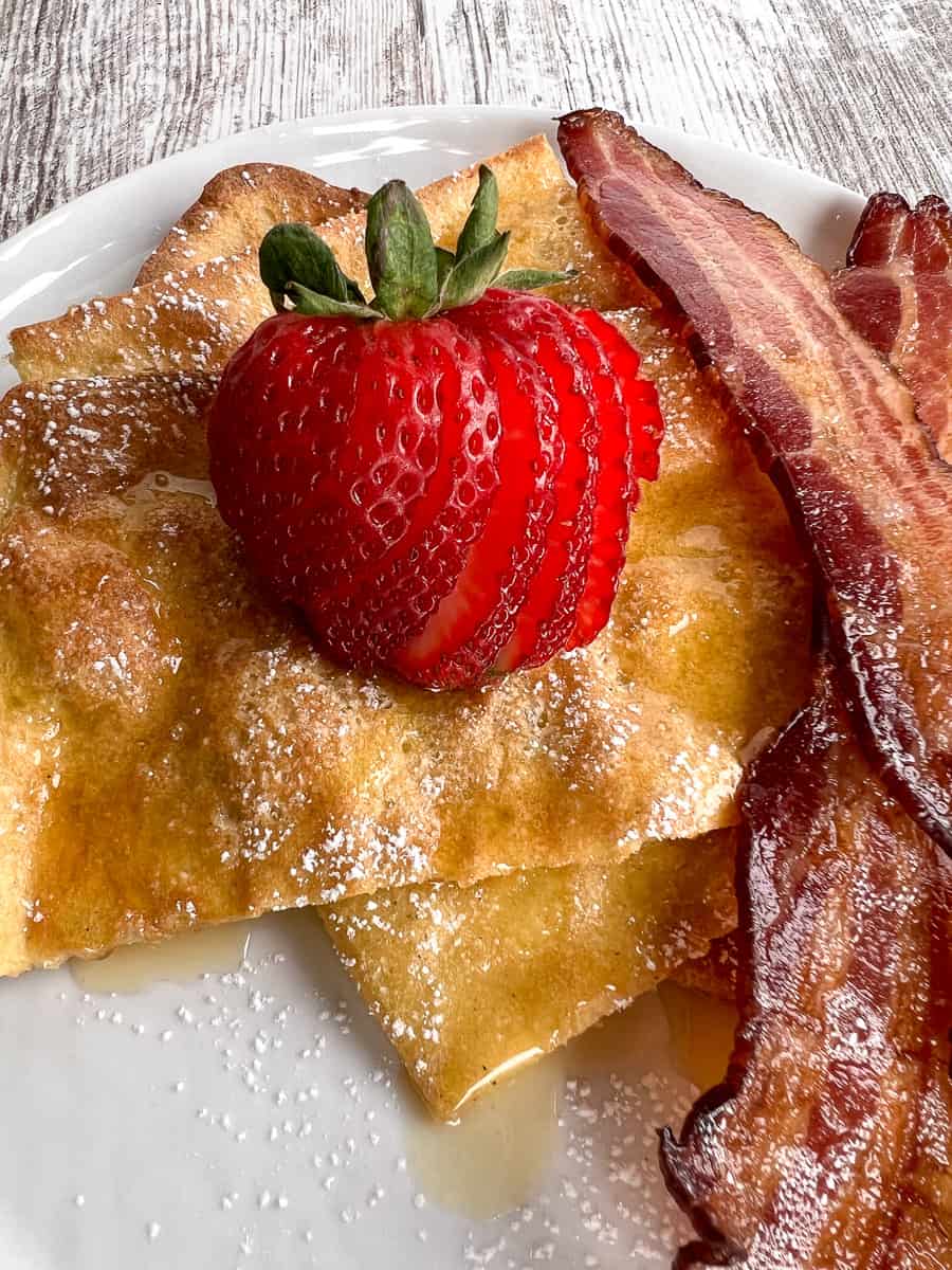pancakes served on a white plate with a side of bacon and sliced strawberry.