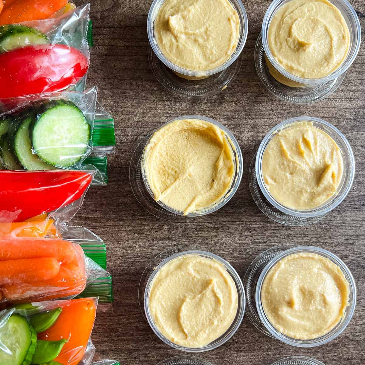 small plastic cups filled with hummus alongside chopped vegetables in a zip top bag.