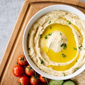 bowl of hummus topped with garlic oil alongside cherry tomatoes on a wooden board.