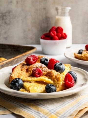 stacked swedish oven pancakes topped with fresh berries alongside a sheet pan of pancakes.