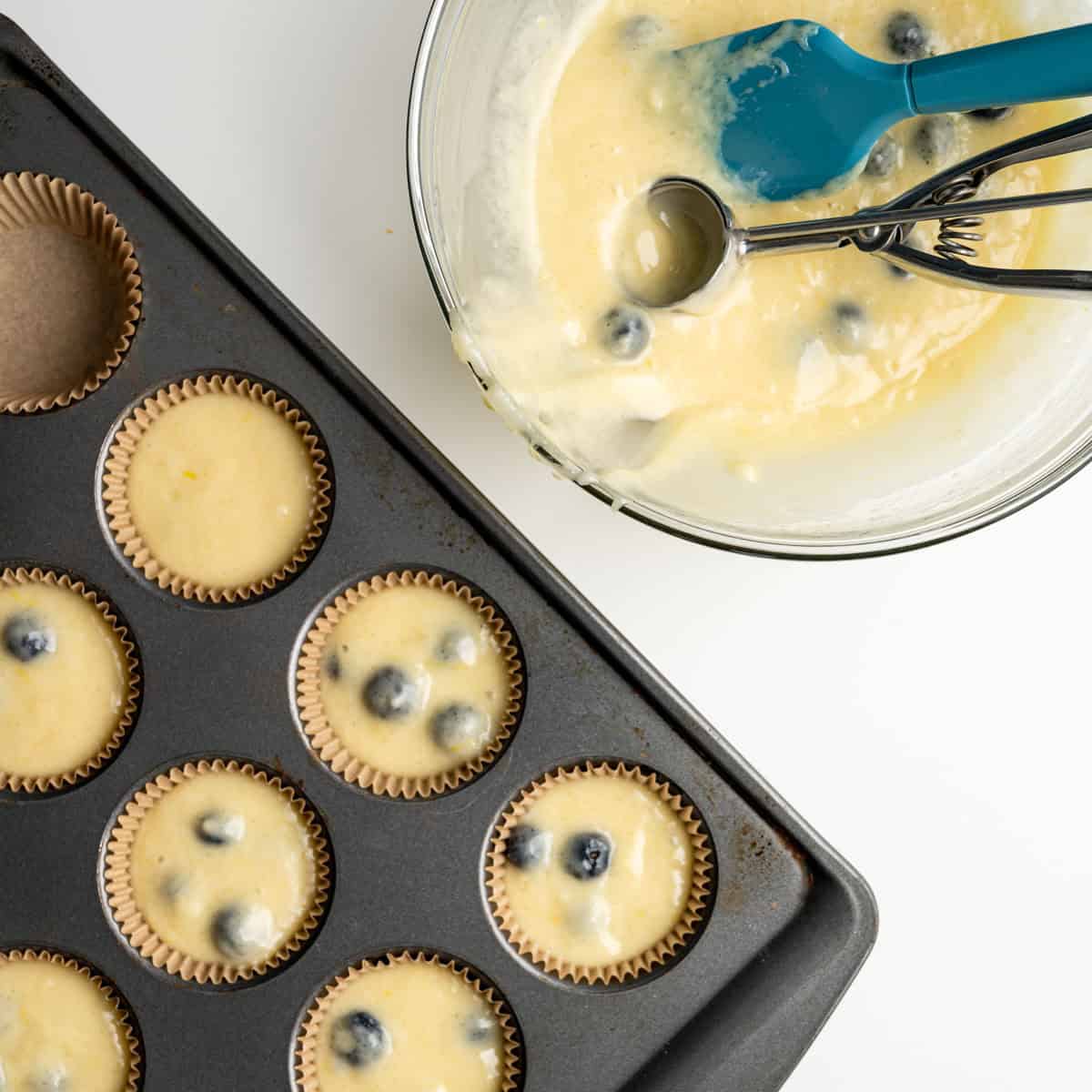 a muffin tin filled with batter alongside a bowl with a cookie scoop and spatula.