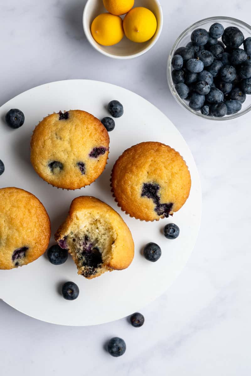 four blueberry muffins on a small white cake stand alongside a bowl of blueberries.
