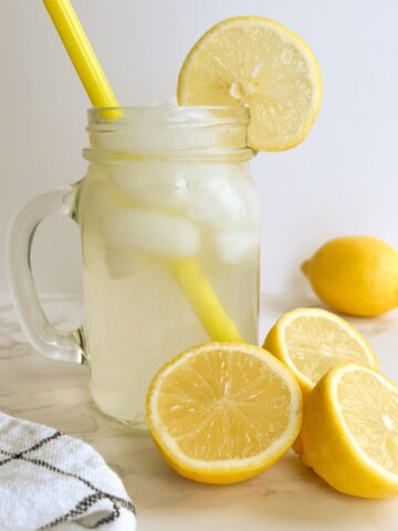 mason jar of lemonade with three sliced lemons in front of the jar, next to a kitchen towel.