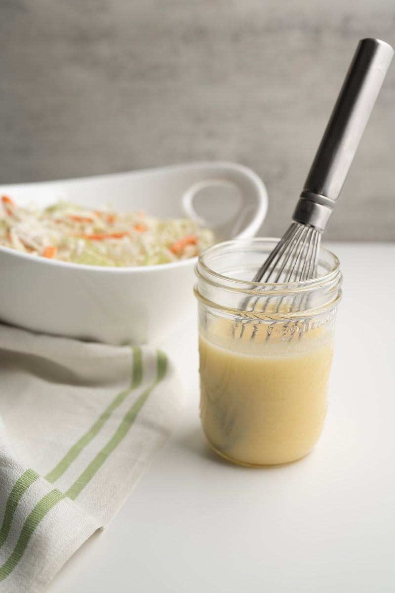 mason jar of coleslaw dressing with a whisk behind it is a white bowl of coleslaw veggies.