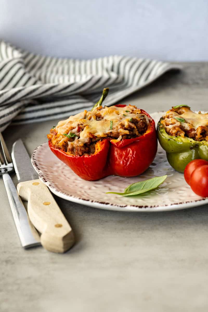 two stuffed peppers on a plate with fresh basil, cherry tomatoes with a Kinfe and fork.