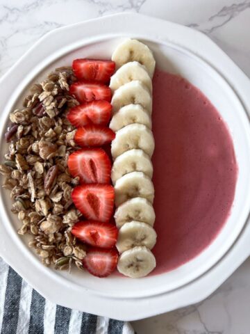 white bowl filled with smoothie topped with banana slices, strawberries and granola.