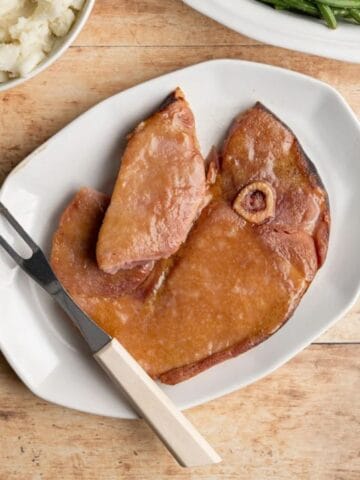 baked ham steak on a platter with a piece cut, with a thick glaze.
