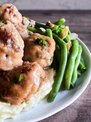 white plate full of swedish meatballs on top of mashed potatoes with a side of green beans.