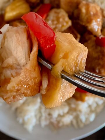 pieces of chicken, red pepper and pineapple on a fork with a white plate in the background.