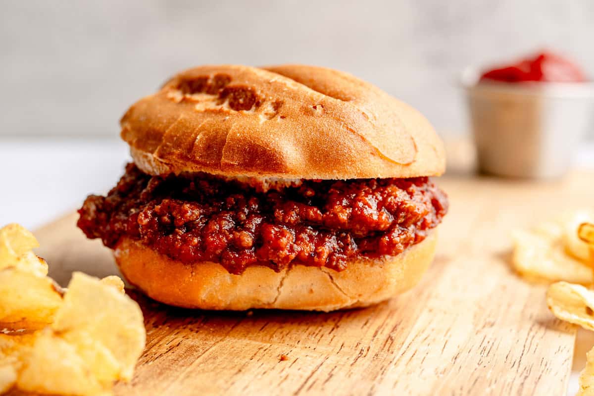 sloppy joe on a wooden board with a handful of chips and a small cup of ketchup.