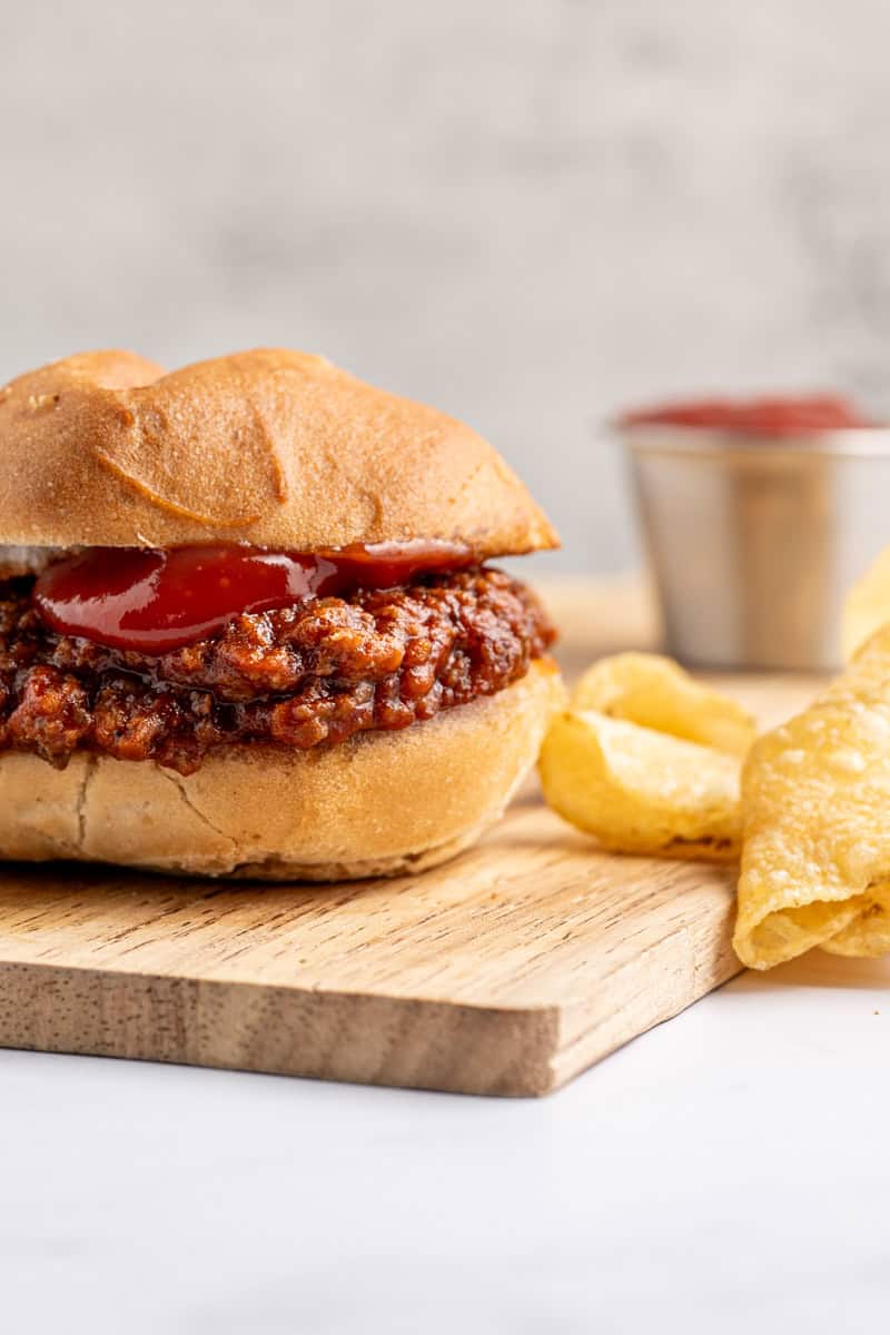 close up of a sloppy joe with ketchup dripping down to a wooden board with a handful of chips and a small dish of ketchup.