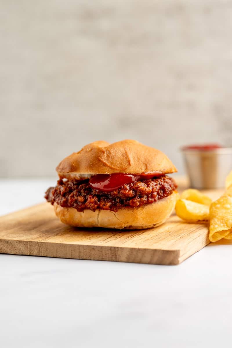 one single sloppy joe on a wooden board with chips and ketchup.