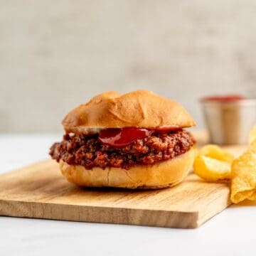 one sloppy joe on a wooden board, alongside a handful of chips and a small dish of ketchup in the background.