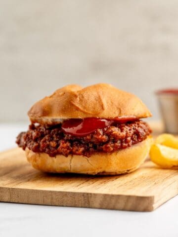 one sloppy joe on a wooden board, alongside a handful of chips and a small dish of ketchup in the background.