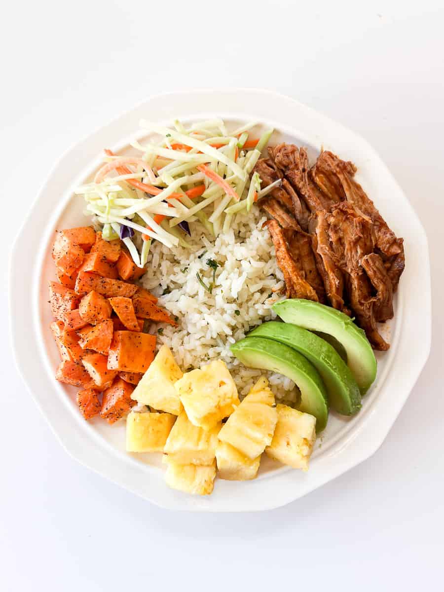white bowl filled with rice, pulled pork, sweet potatoes, broccoli slaw, avocado, and pineapple.