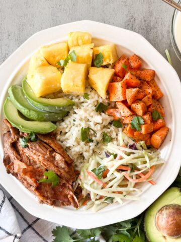a white bowl filled with rice, pulled pork, sweet potatoes, slaw, pineapples and avocados.