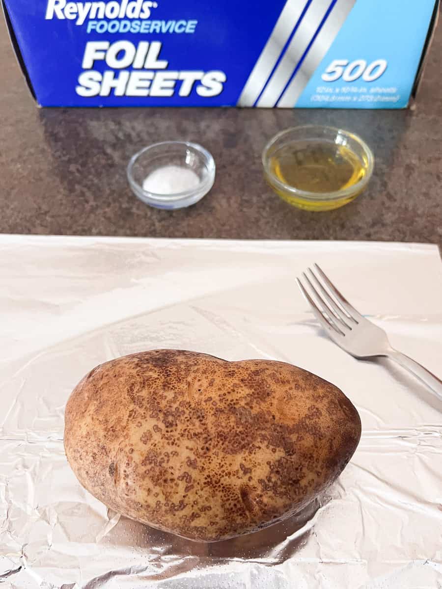 potato in the center of tinfoil with a small bowl of salt and oil with a fork. 