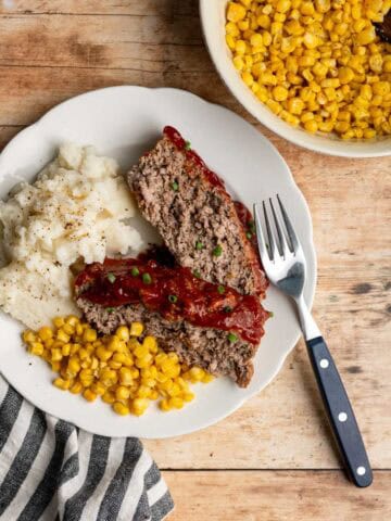 dinner plat with two slices of meatloaf, a scoop of mashed potatoes and sweet corn on a wooden table.