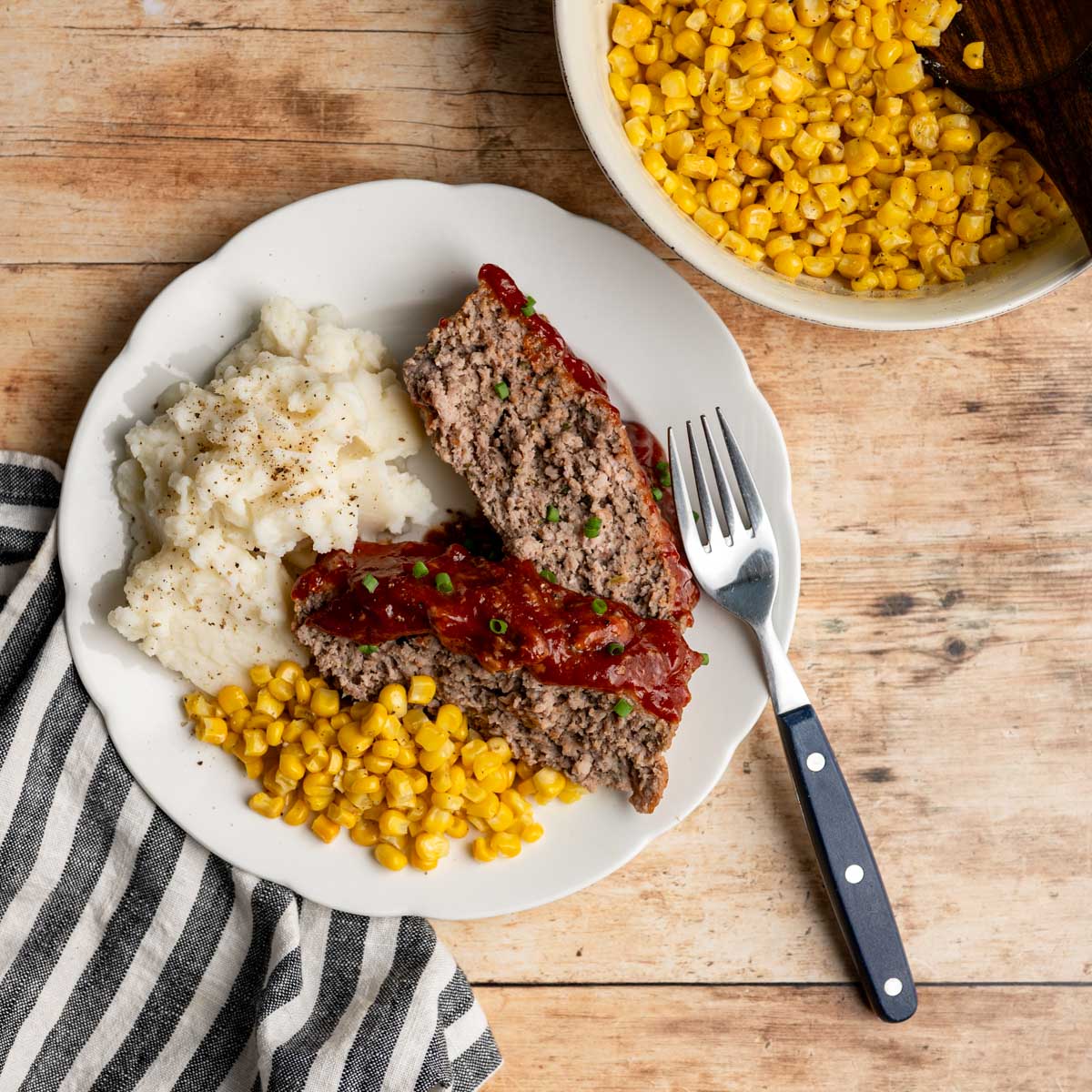 dinner plate with two slices of meatloaf, mashed potatoes and corn on a wooden table.