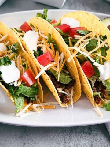 three tacos on a white platter topped with lettuce, tomatoes, cheese and sour cream.