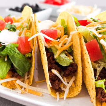 three hard shell tacos with toppings in the background.