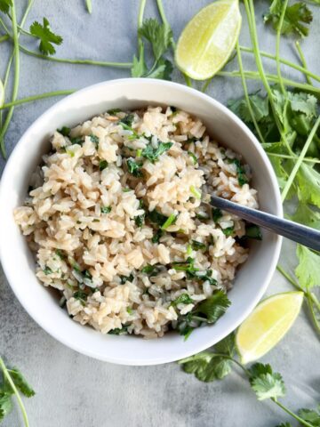 white bowl with rice and a spoon surrounded by cilantro and lime wedges.