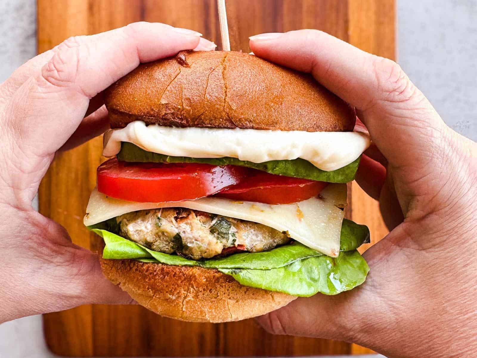 two hands holding a chicken burger over a wooden cutting board.