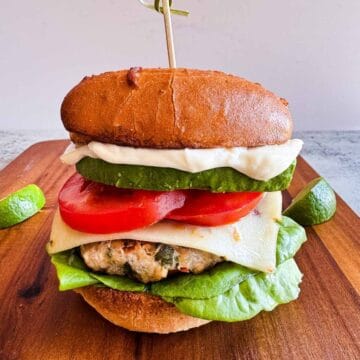 ground chicken burger on a gluten free bun, stacked with avocado, tomato, pepper jack cheese and lettuce on a wooden cutting board.