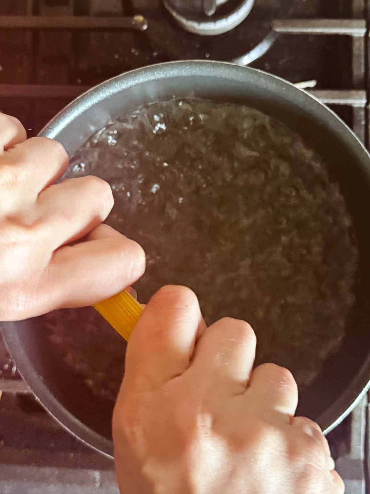 two hands breaking spaghetti noodles over a pot of boiling water.