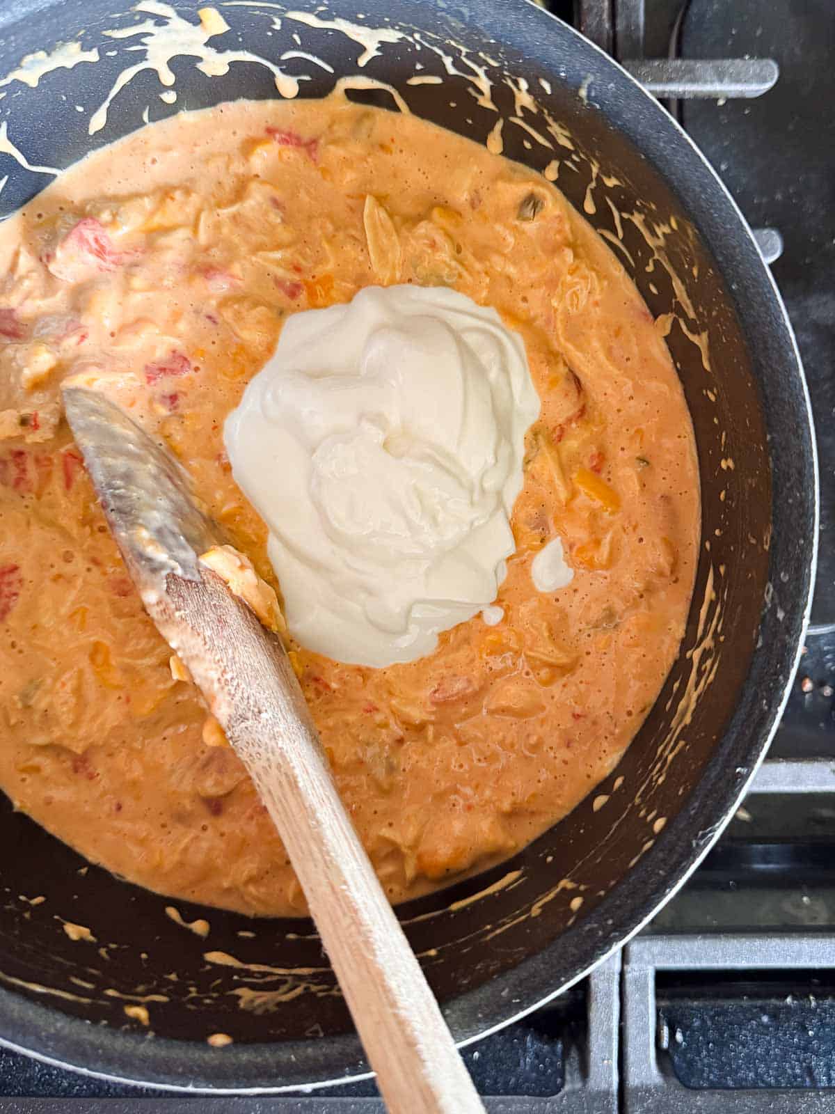saucepan on stovetop with cheesy sauce and a scoop of sour cream with a wooden spoon.