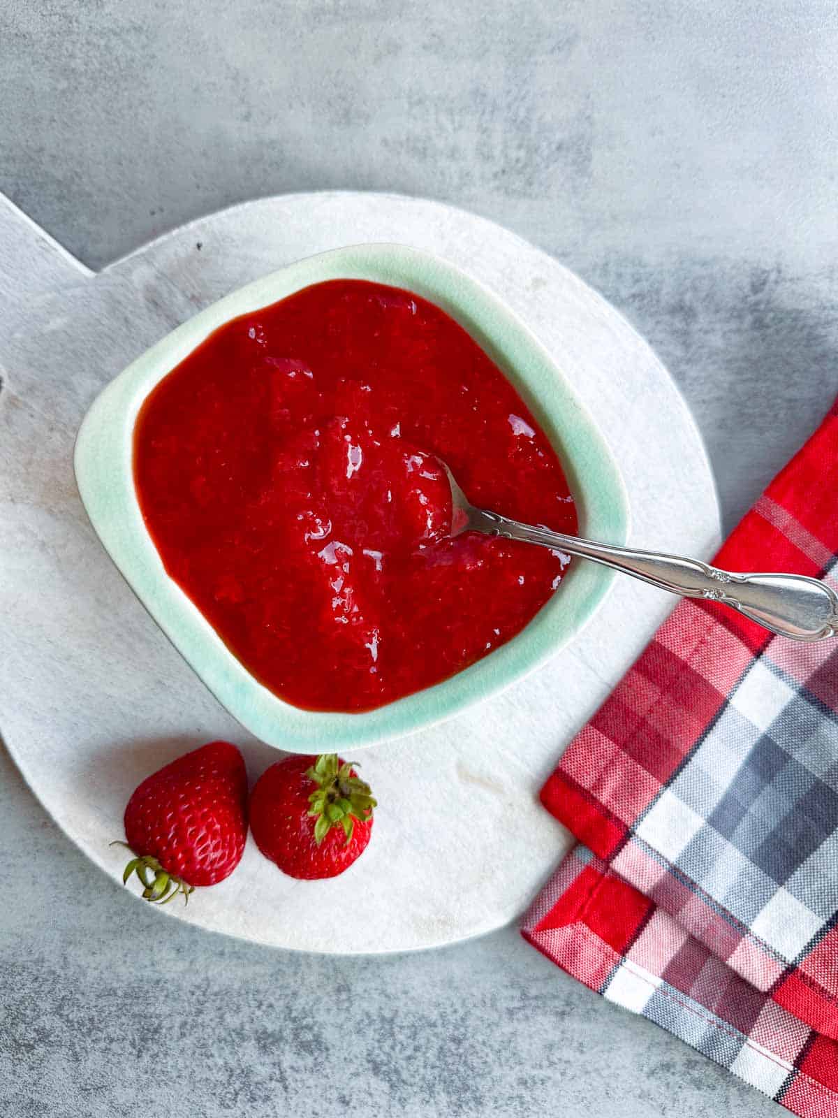 mint green bowl of strawberry sauce beside two whole berries.
