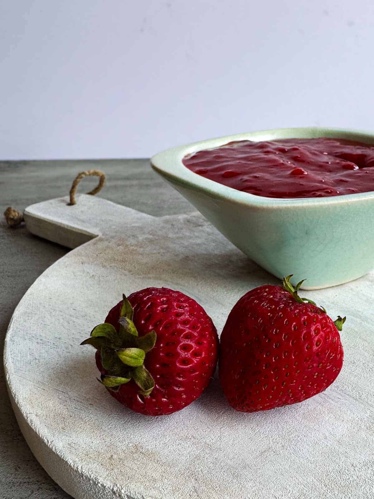 small green bowl of strawberry sauce behind two whole strawberries on a white wooden board.