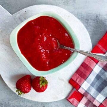 mint green bowl of strawberry sauce beside two whole berries.