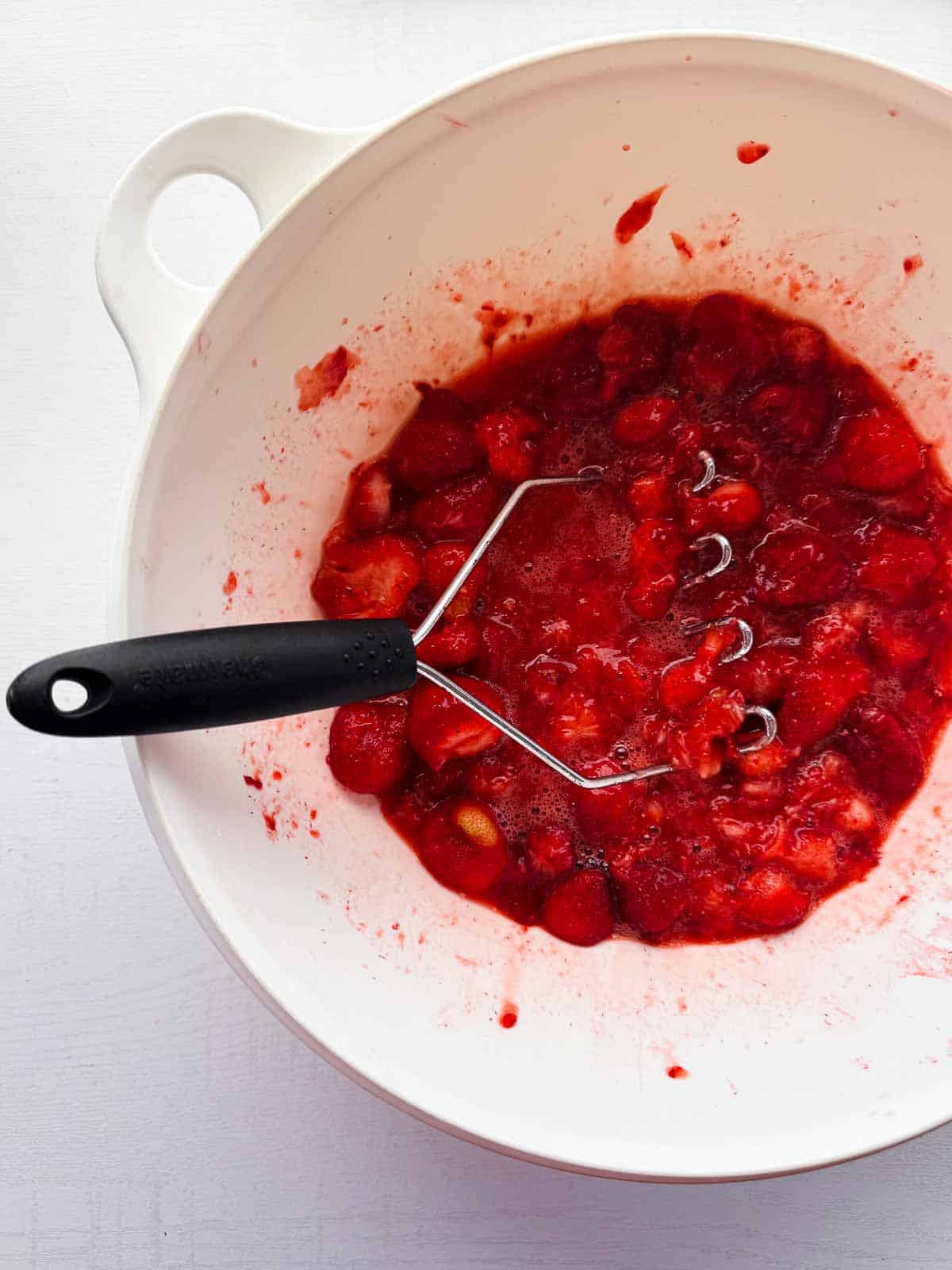 large white bowl with crushed strawberries and a potato masher.