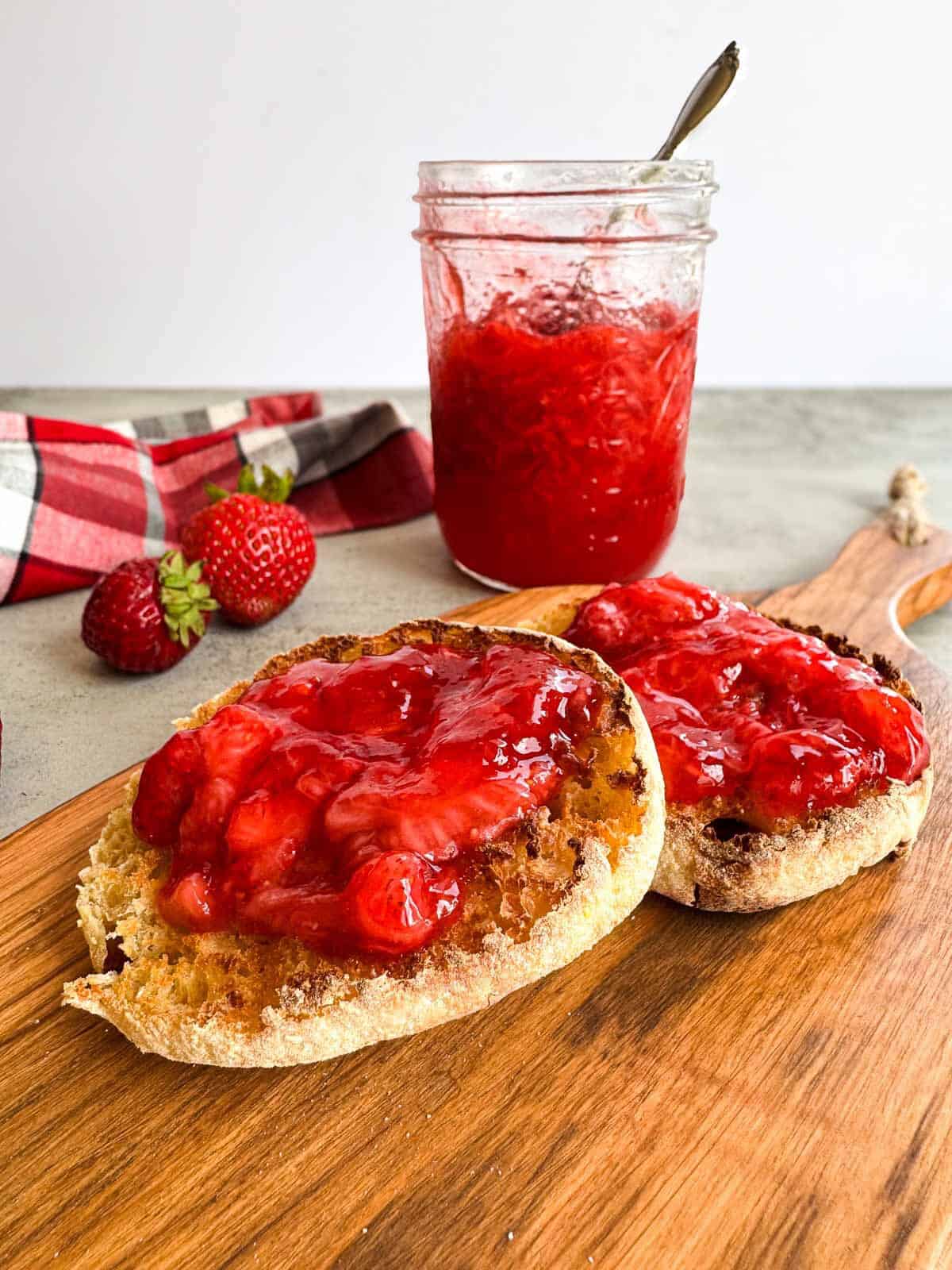 english muffin topped with strawberry jam on a wooden cutting board with a jar in the background.