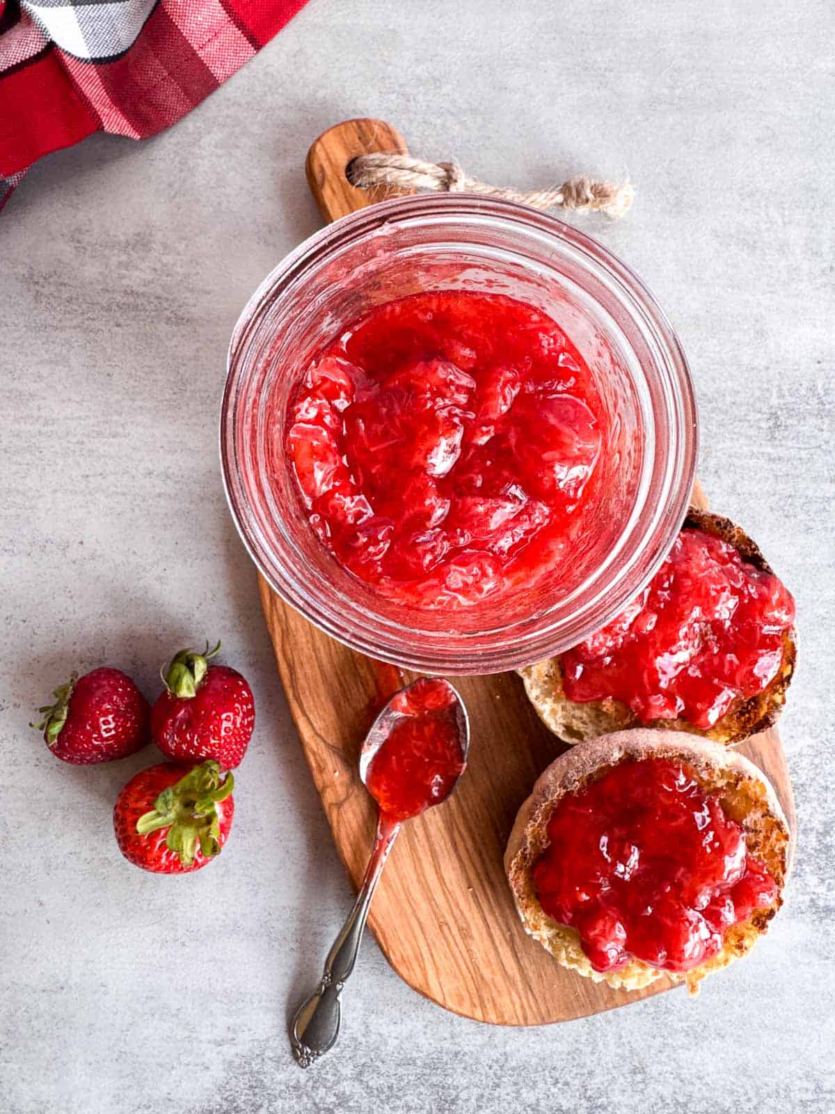 flat lay with an open jar of strawberry jam with a toasted english muffin and a spoonful on a wooden cutting board. 