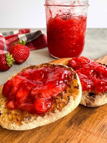 toasted english muffin topped with strawberry jam on a wooden cutting board, a jar of strawberry jam with three whole strawberries.