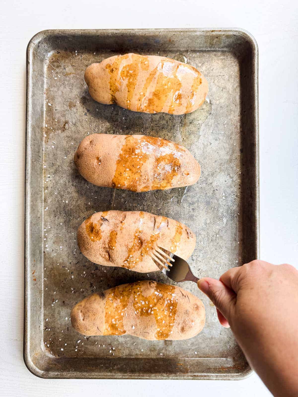 four potatoes on a baking sheet drizzled with avocado oil, salt and being poked with a fork.