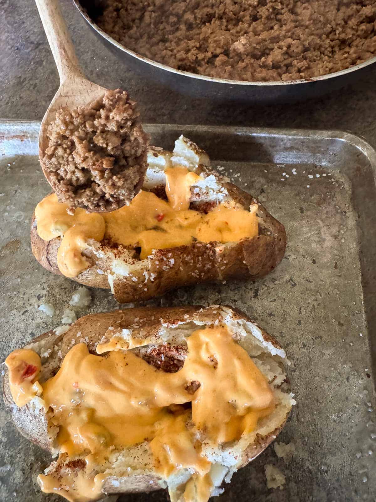 two baked potatoes topped with queso and a small wooden spoonful of taco flavored beef into the potato.