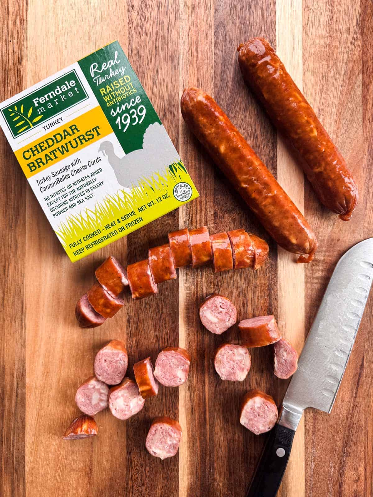 two turkey brats sliced and two whole turkey brats on a wooden cutting board alongside a knife.