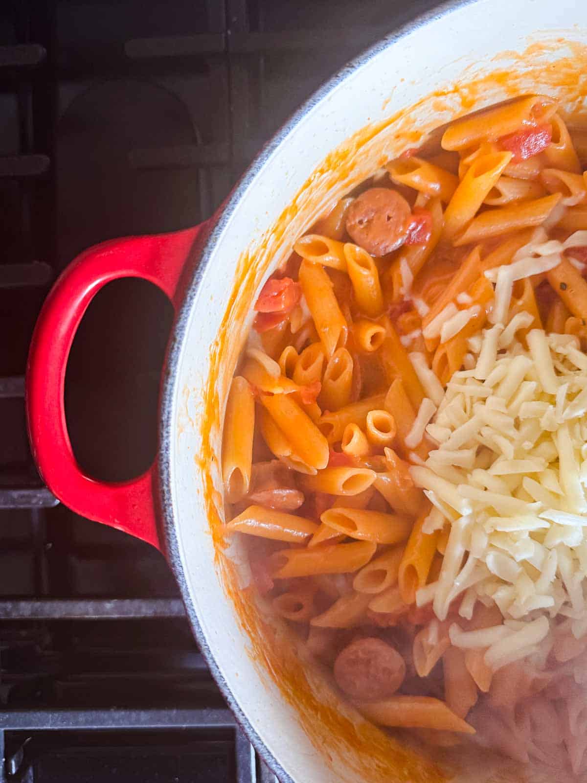 cooked penne pasta in a red dutch oven topped with shredded cheese.