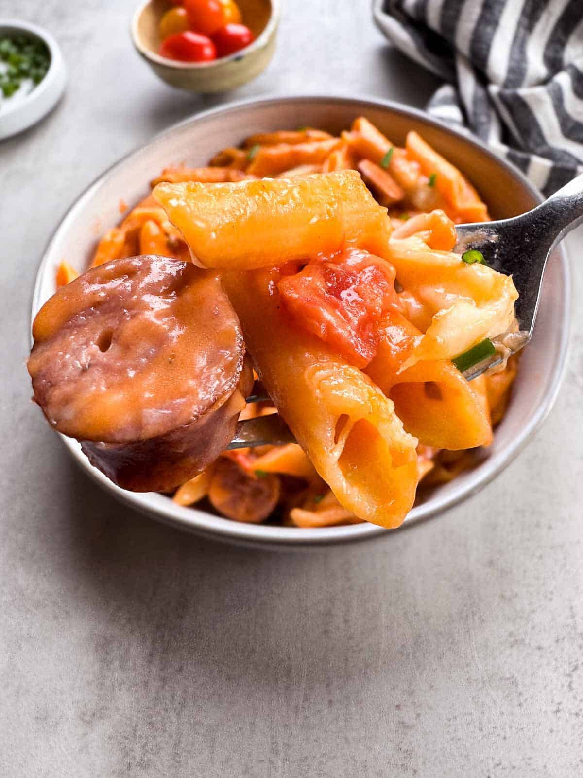 fork full of sliced turkey brat, penne pasta, diced tomato and a cheese curds above a bowl of pasta.