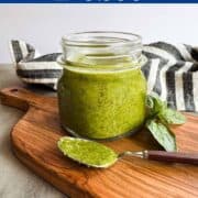 Glass jar of fresh green low FODMAP pesto on a wooden cutting board with a spoonful of pesto in front and basil leaves beside it.