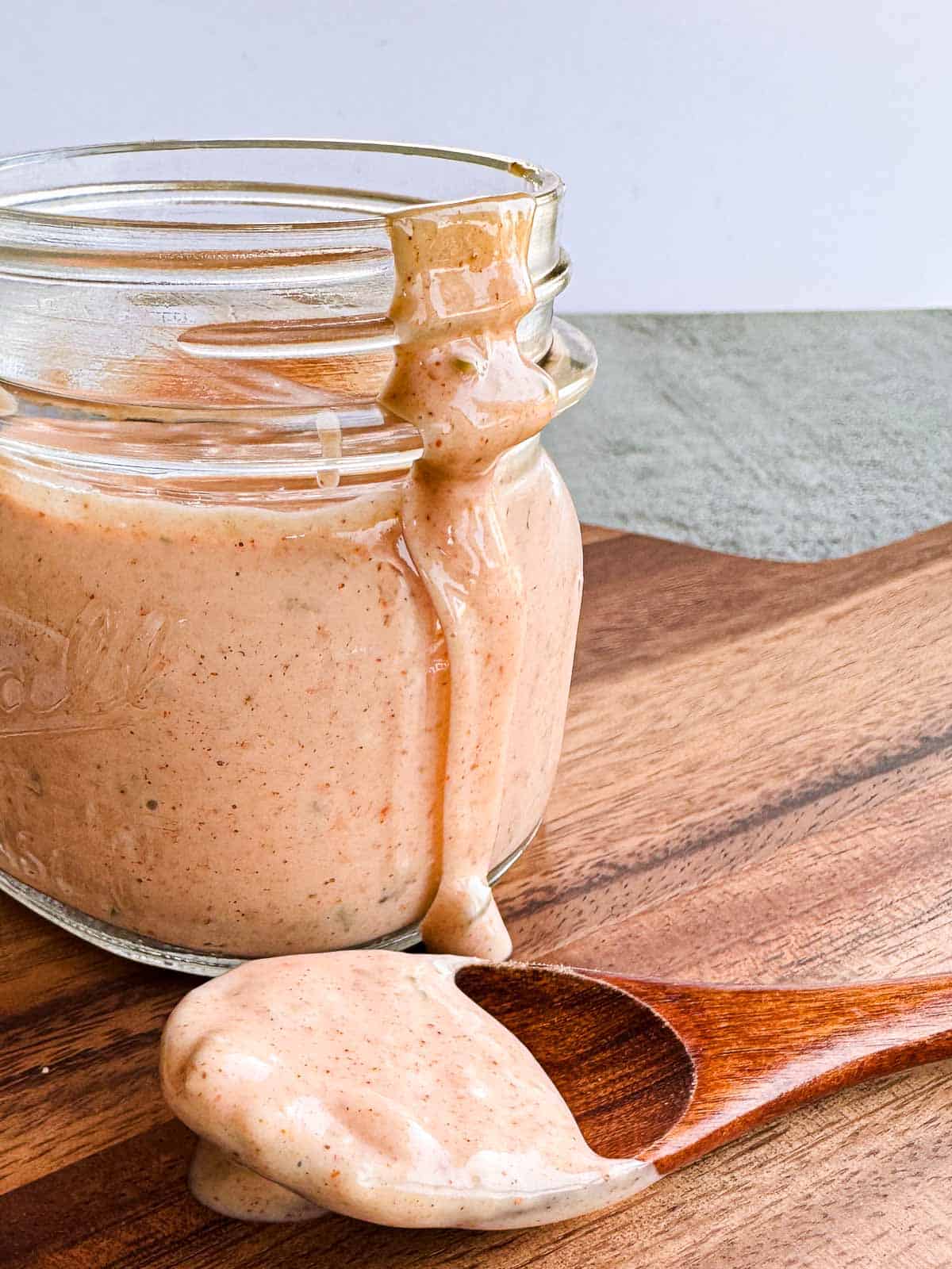 small mason jar sitting on a wooden board alongside a small wooden spoon filled with thousand island dressing and a napkin.