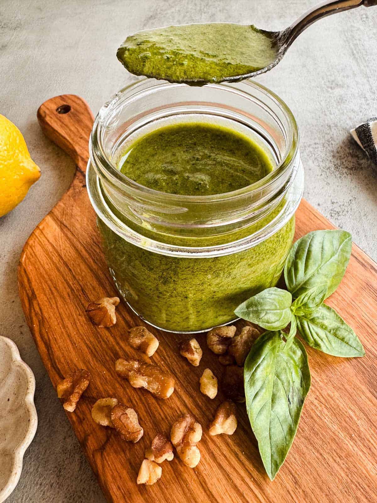 mason jar with a spoonful of pesto on a wooden cutting board with fresh basil, walnuts, lemon and salt.
